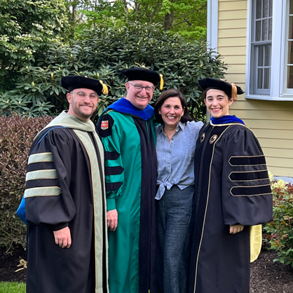 Aaron S. Marks ’24 DHSc with his father, Brian, mother, Lynne, and sister, Madeline. The family stands in front of a yellow house and garden, arms around each other, looking forward, all in their cap and gowns.